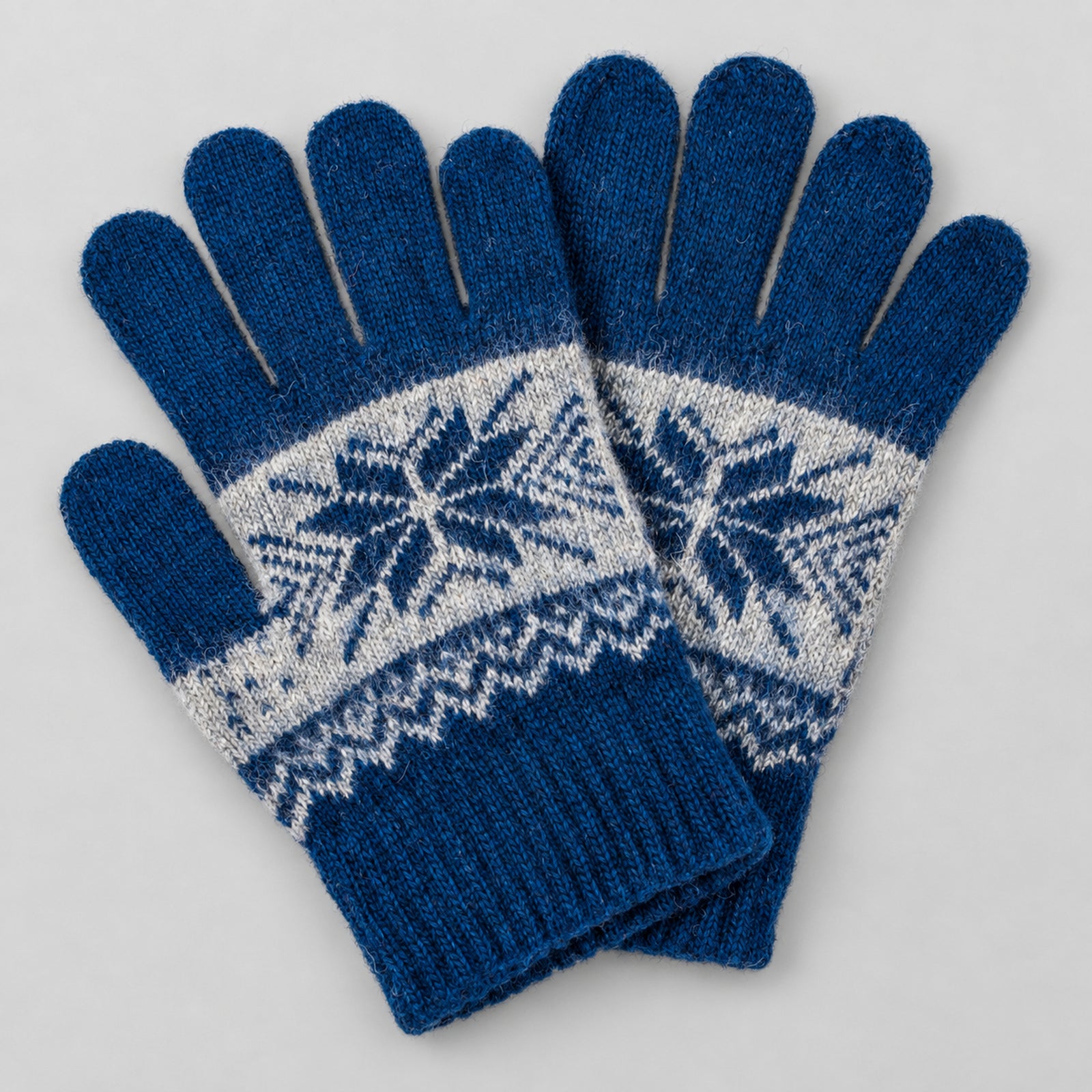 A pair of blue knit winter gloves with a white Fair Isle snowflake pattern across the back of the hands, displayed on a light gray background.