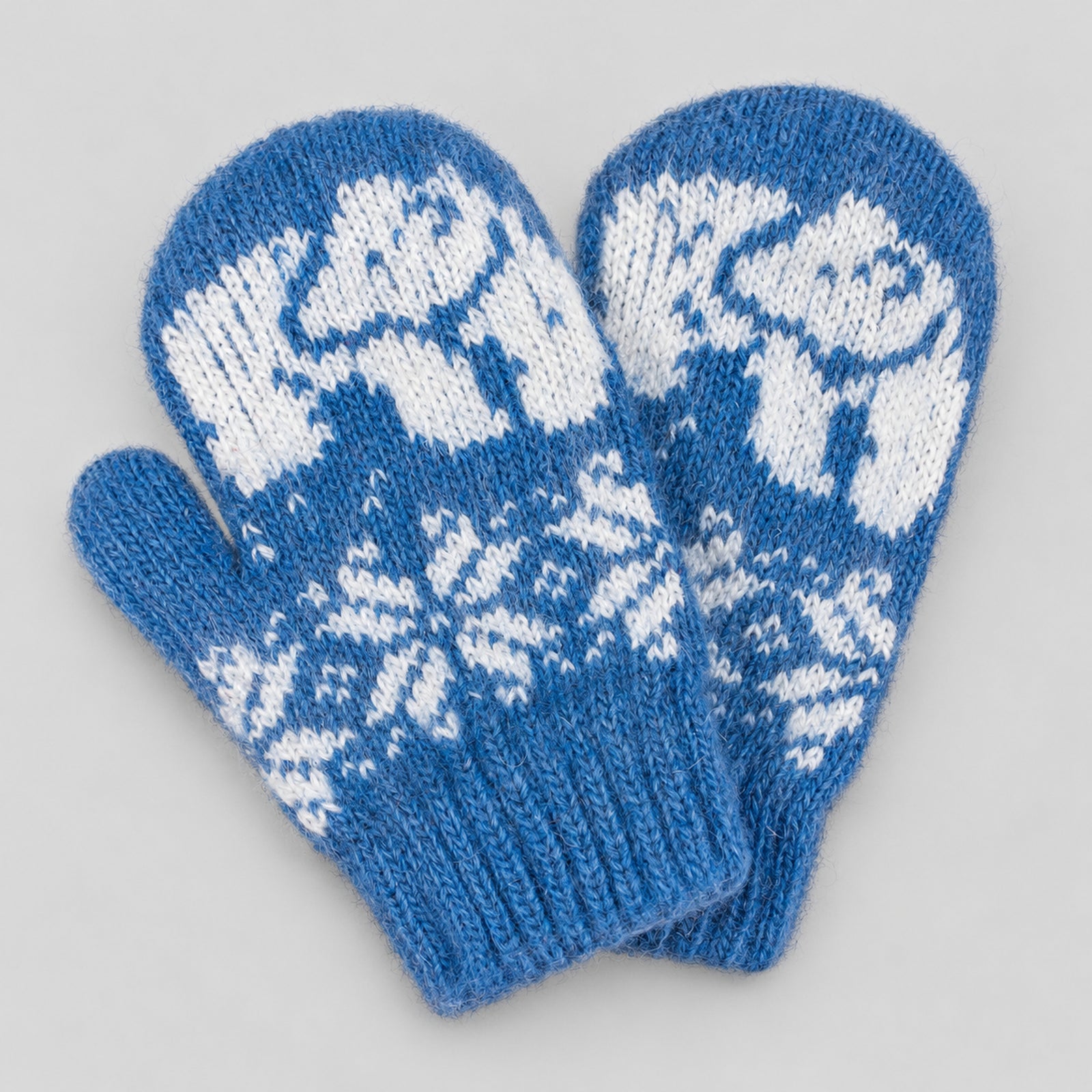 A pair of blue knit mittens with white fair isle patterns featuring polar bears and snowflakes, displayed on a neutral light gray background.