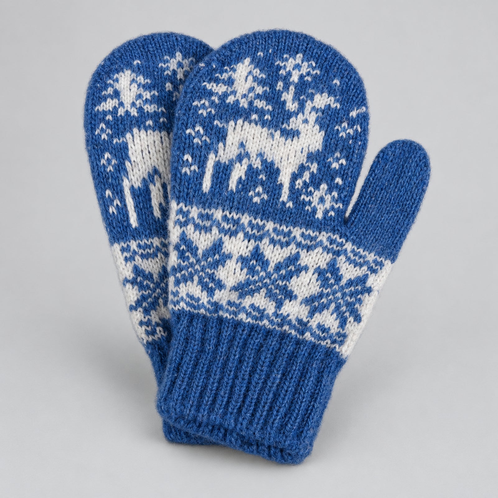 A pair of blue and white knit winter mittens featuring a fair isle pattern with reindeer and snowflakes, displayed on a neutral light gray background.