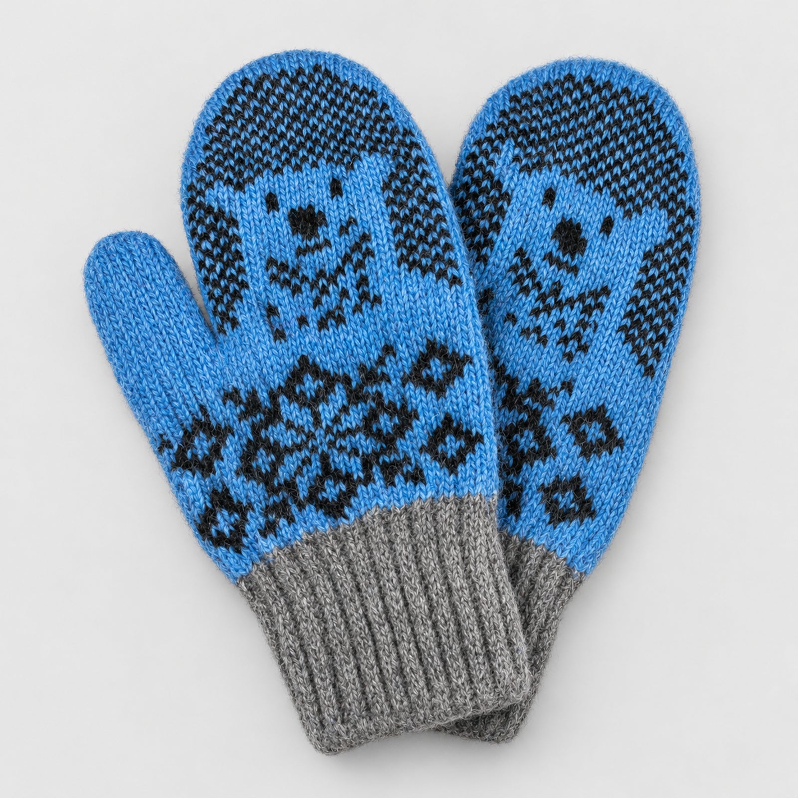 A pair of knit winter mittens in bright blue and gray with black dog face and diamond patterns, shown on a light gray background.