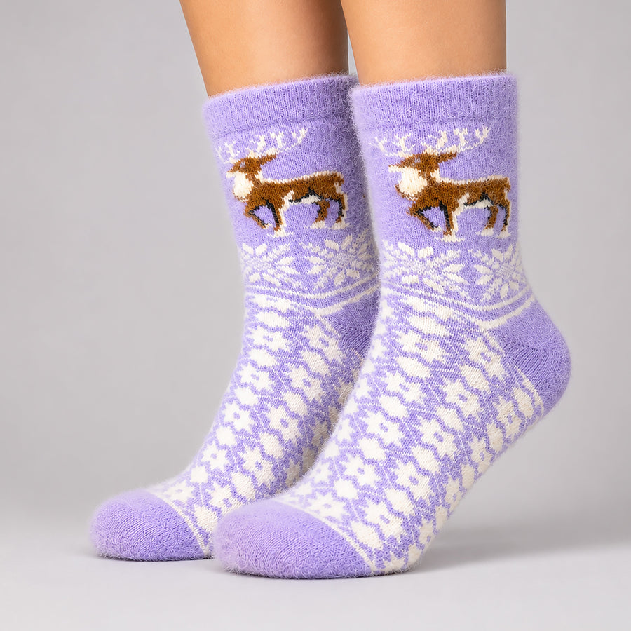 A pair of fuzzy crew socks worn on feet, featuring a light purple base with a white Fair Isle snowflake pattern and brown reindeer motifs on the upper leg.