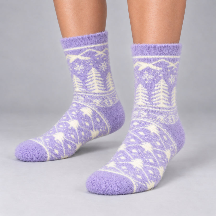A pair of fuzzy crew socks in lavender purple and cream, featuring a Nordic-style pattern of pine trees and snowflakes, shown on a person's feet against a neutral gray background.