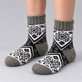 A pair of crew socks with a black, white, and gray Nordic snowflake pattern, shown on a person's feet against a light gray studio background.