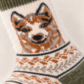 Close-up of a knitted sock featuring a dog face in orange, cream, and black, with a patterned knit body and dark green cuff.