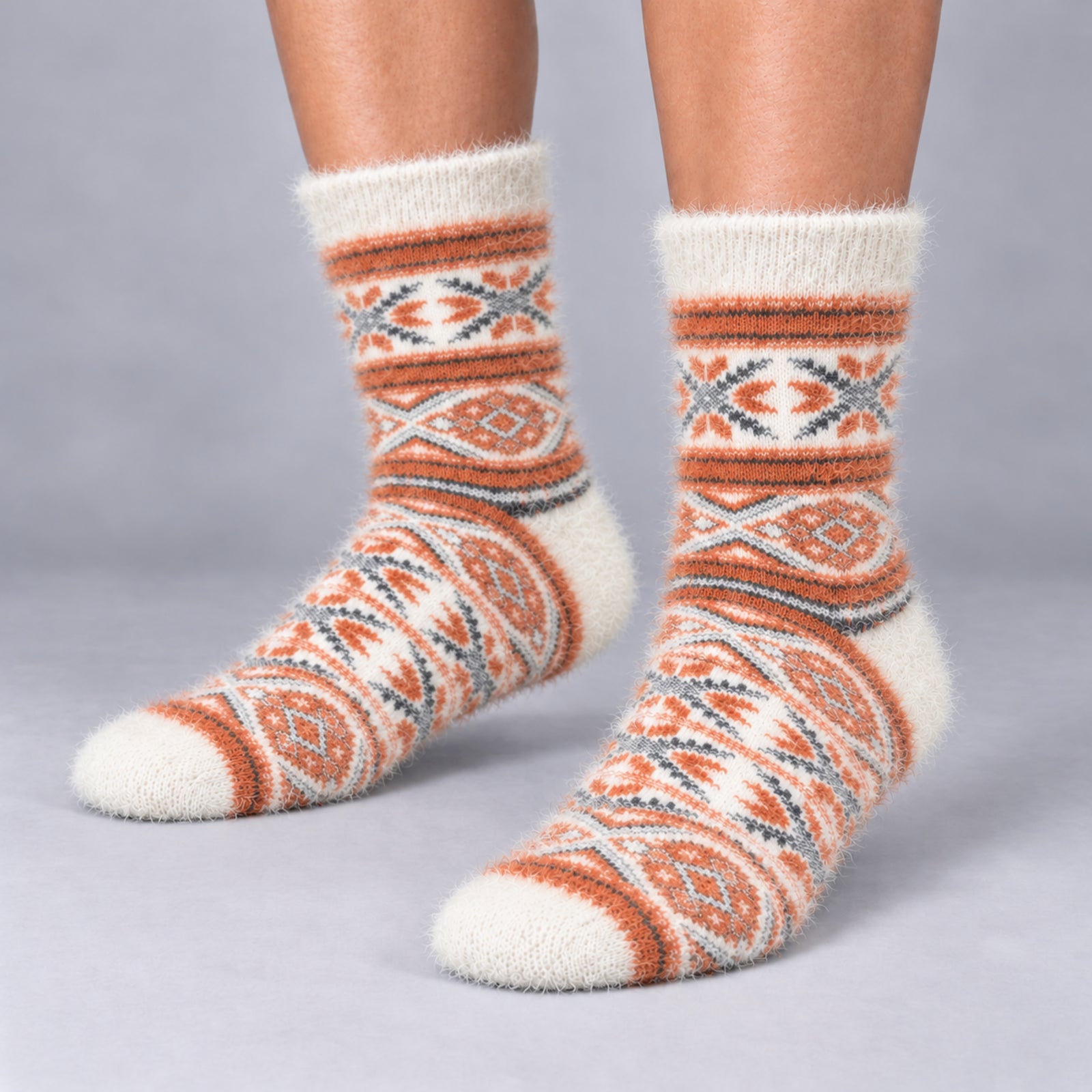 A pair of fuzzy crew socks with a Southwestern geometric pattern in orange, cream, and gray, shown on a person's feet against a neutral gray background.