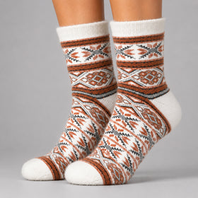 A pair of crew socks with a Southwestern geometric pattern in rust orange, gray, and white, shown on a person's feet against a solid gray studio background.