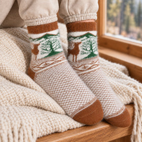 A close-up of a person's feet wearing cream knit socks with a brown and green forest and deer pattern, resting on a cozy cream knit blanket near a wooden window.