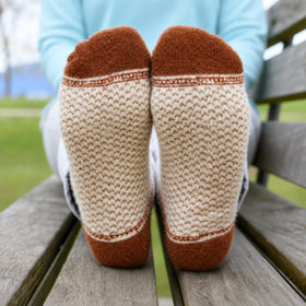 A close-up of a person sitting on a wooden bench outdoors, showing the soles of their feet wearing cream and rust-colored knit socks.