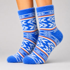 A pair of fuzzy blue socks with white and orange geometric patterns, worn on a person's feet against a light gray studio background.