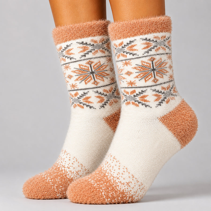 A pair of fuzzy crew socks worn on feet, featuring a cream base with orange and gray Fair Isle style snowflake patterns on the leg and a solid orange toe and heel sections.