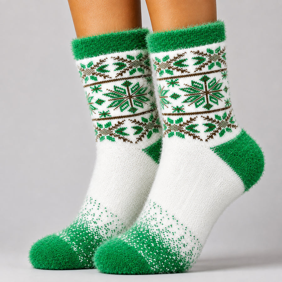 A pair of fuzzy crew socks worn on a person's feet, featuring a white base with green and brown Nordic-style snowflake and geometric patterns on the upper leg, and solid green heels, toes, and cuffs.