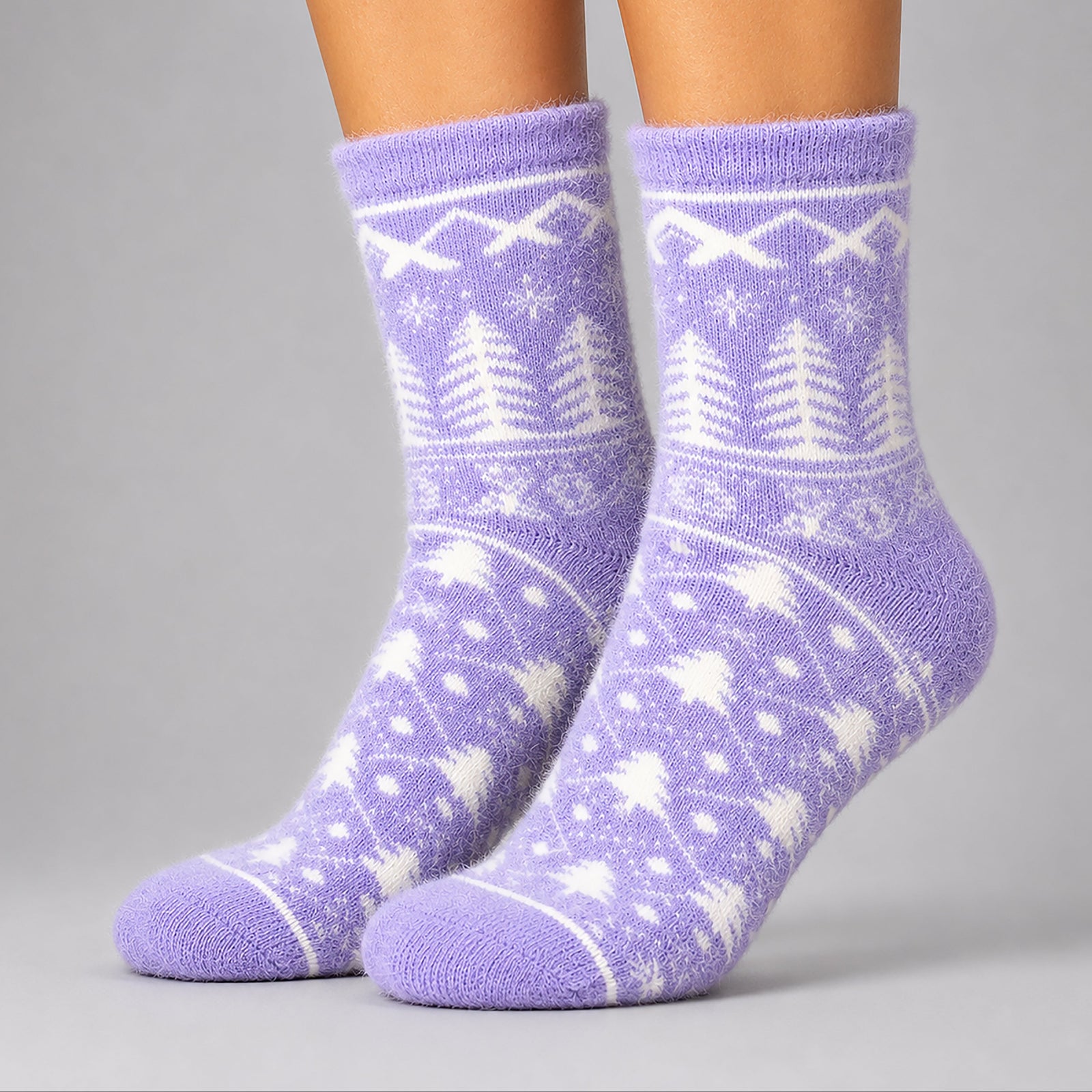 A pair of fuzzy crew socks worn on feet, featuring a white Nordic-style pattern of trees and snowflakes on a light purple background, photographed against a neutral gray studio backdrop.