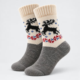 A pair of crew socks with a gray knit foot and leg section, a cream-colored upper cuff and leg section featuring a black reindeer and red and blue Nordic-style pattern, and ribbed cream cuffs.