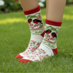 A pair of white knit socks with red and green holiday patterns, featuring two black birds perched on branches, worn on a person's feet on a grass.