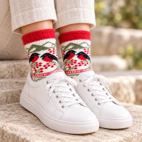 A close-up of a person's feet wearing white leather sneakers and cream-colored trousers, showcasing a pair of festive knit crew socks with a red, green, and black bird pattern.