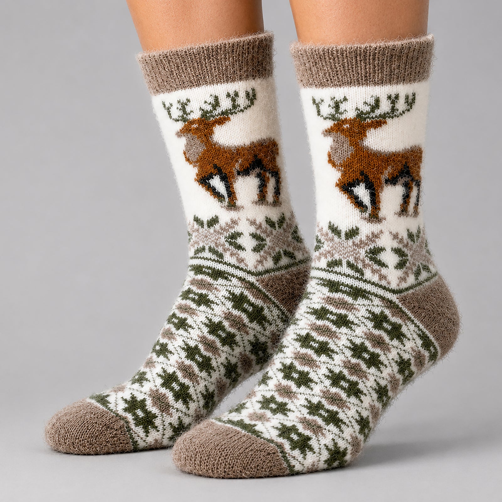 A pair of crew socks worn on feet, featuring a white background with a brown reindeer graphic on the upper leg, green snowflake patterns, and brown knit cuffs, heels, and toes on a light gray studio background.