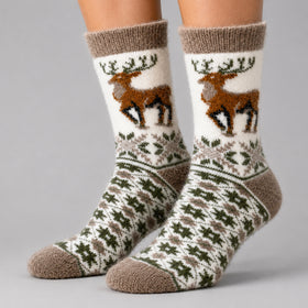 A pair of crew socks worn on feet, featuring a white background with a brown reindeer graphic on the upper leg, green snowflake patterns, and brown knit cuffs, heels, and toes on a light gray studio background.