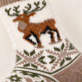 A close-up of a knitted Christmas sock featuring a brown reindeer with black and white details, green antlers, and a green and brown geometric pattern on a cream background.