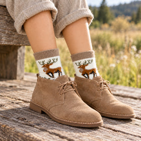 A person sitting outdoors on a wooden bench wearing tan suede chukka boots and cream knit socks with a brown deer pattern.