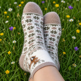 A pair of patterned knit socks worn on feet resting on green grass with small yellow and white flowers. The socks have a cream base with brown and olive green Fair Isle style motifs and a small brown animal design near the ankle.