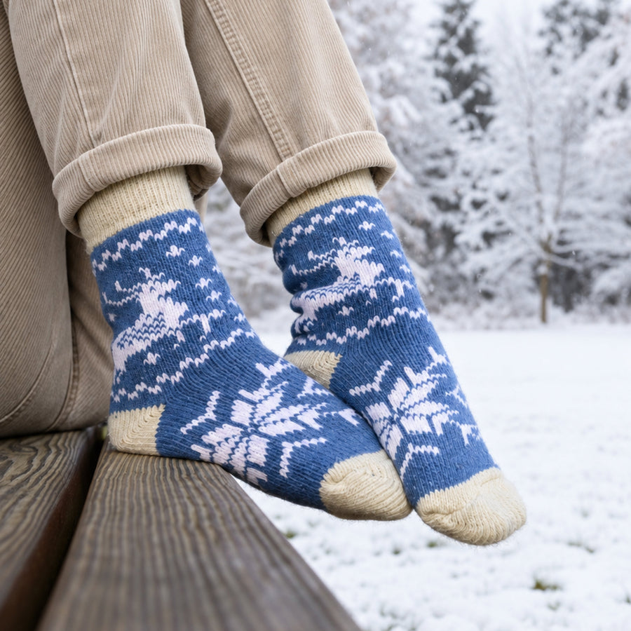 A person sitting outdoors in the snow wears blue and cream knit socks with white snowflake and reindeer patterns, paired with beige corduroy pants.