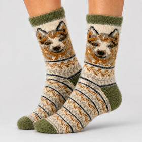 A pair of crew socks worn on feet, featuring a knitted dog face design in brown, cream, and black, with olive green cuffs, heels, and toes, set against a light gray studio background.