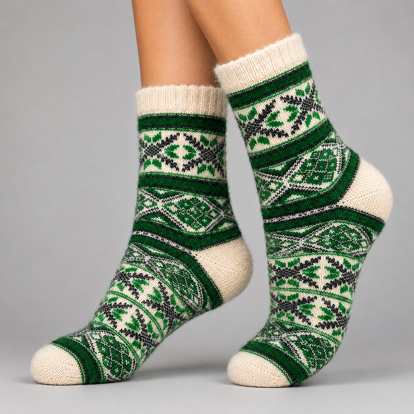 A pair of cream-colored knit socks with a green and black Nordic/Fair Isle pattern, worn on a person's feet against a solid gray studio background.