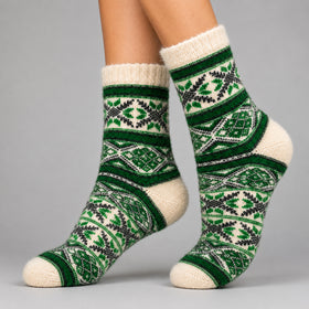 A pair of cream-colored knit socks with a green and black Nordic/Fair Isle pattern, worn on a person's feet against a solid gray studio background.