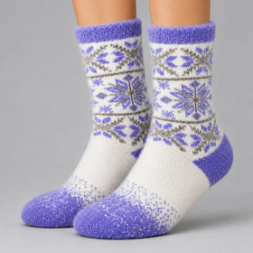 A pair of fuzzy crew socks worn on feet, featuring a white base with purple and gray Nordic snowflake patterns and solid purple toes, heels, and cuffs, set against a neutral gray background.