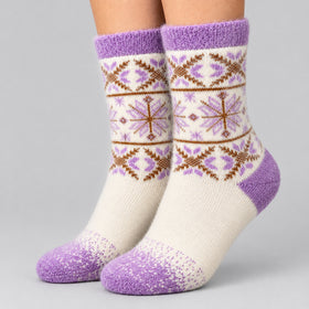 A pair of crew socks worn on feet, featuring a cream base with purple and brown Fair Isle snowflake and geometric patterns, set against a solid light gray background.