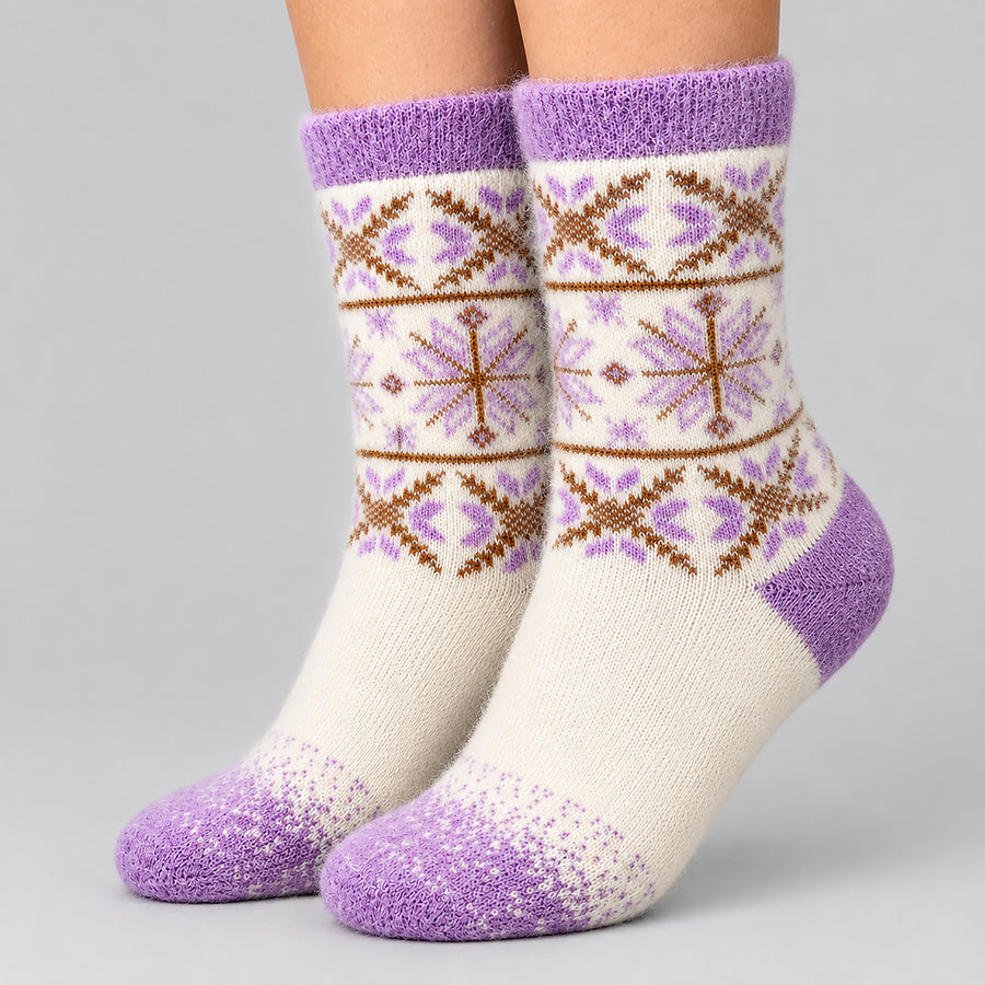 A pair of crew socks worn on feet, featuring a cream base with purple and brown Fair Isle snowflake and geometric patterns, set against a solid light gray background.