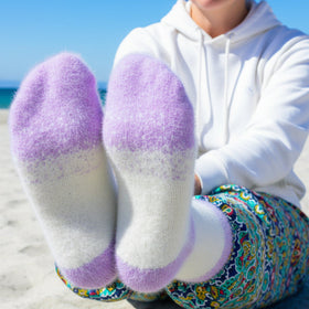 Person wearing white socks with purple toes sitting on a beach.