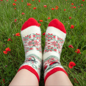 A person's legs and feet wearing white knit socks with red toes, heels, and cuffs, decorated with a red and green floral pattern, sitting in a field of red poppies.