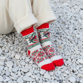 A pair of white knit socks with red and green fair isle patterns and bird motifs, worn with blue jeans on a pebble beach.