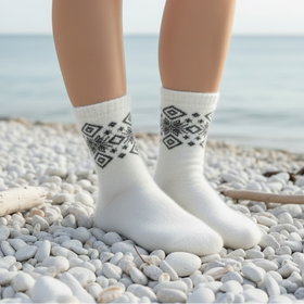 A pair of white fuzzy crew socks with a black geometric snowflake pattern around the cuffs, shown on a person's feet standing on a pebble beach.