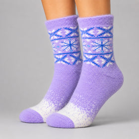 A pair of fuzzy crew socks worn on feet, featuring a lavender purple body with a blue and white Fair Isle snowflake pattern around the calf and a white speckled gradient near the toes, set against a solid gray studio background.