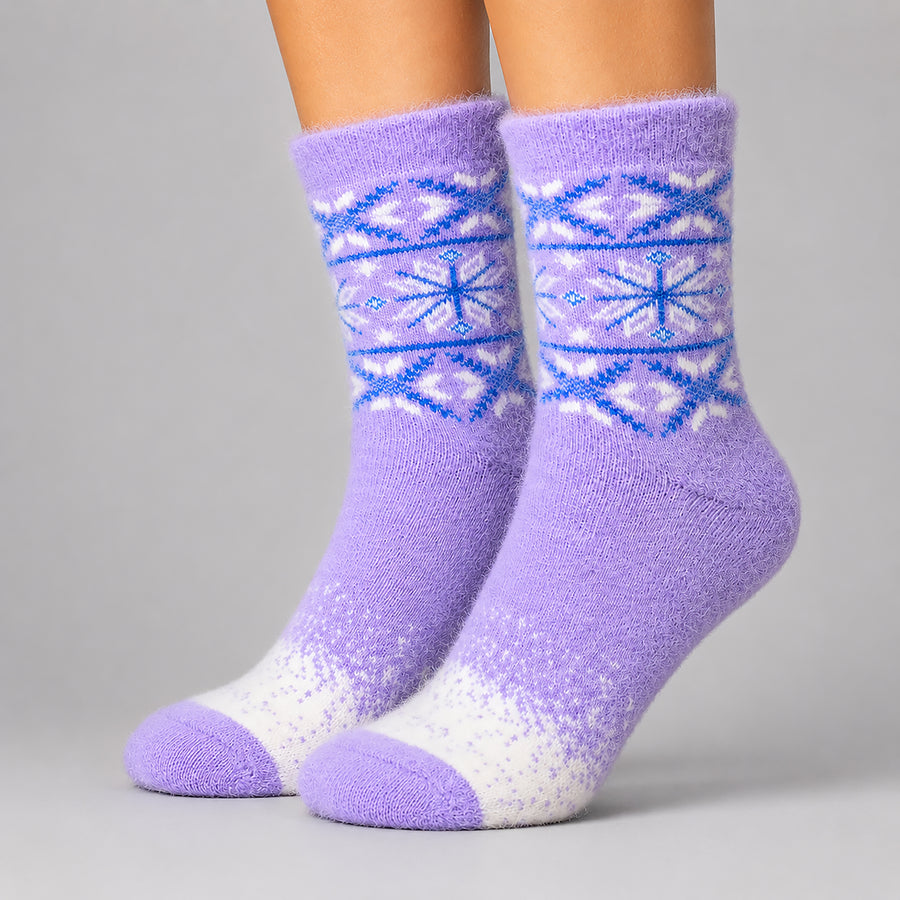 A pair of fuzzy crew socks worn on feet, featuring a lavender purple body with a blue and white Fair Isle snowflake pattern around the calf and a white speckled gradient near the toes, set against a solid gray studio background.