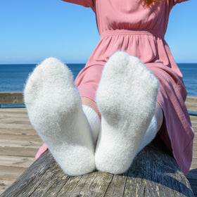 Person wearing white fuzzy socks sitting on a wooden bench by the ocean.