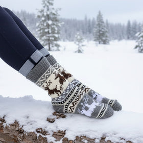 Person wearing patterned socks sitting on a log in a snowy landscape with trees in the background.